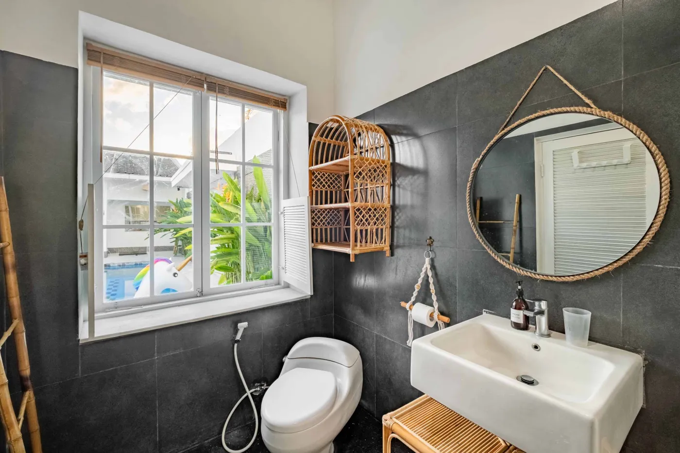 Modern tropical bathroom with dark tiled walls, rattan shelf and bamboo accents, oval rope mirror, white sink, bidet toilet, and window view of poolside greenery. - Bali Villas