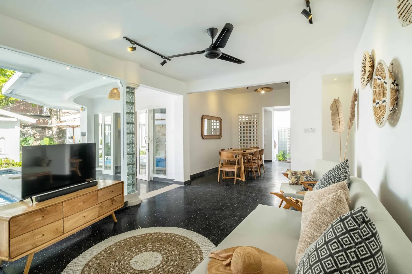 Modern tropical bathroom with dark tiled walls, rattan shelf and bamboo accents, oval rope mirror, white sink, bidet toilet, and window view of poolside greenery. - Bali Villas