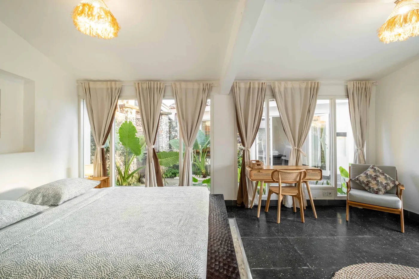 Modern tropical bathroom with dark tiled walls, rattan shelf and bamboo accents, oval rope mirror, white sink, bidet toilet, and window view of poolside greenery. - Bali Villas