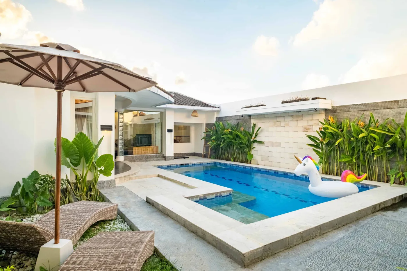 Modern tropical bathroom with dark tiled walls, rattan shelf and bamboo accents, oval rope mirror, white sink, bidet toilet, and window view of poolside greenery. - Bali Villas