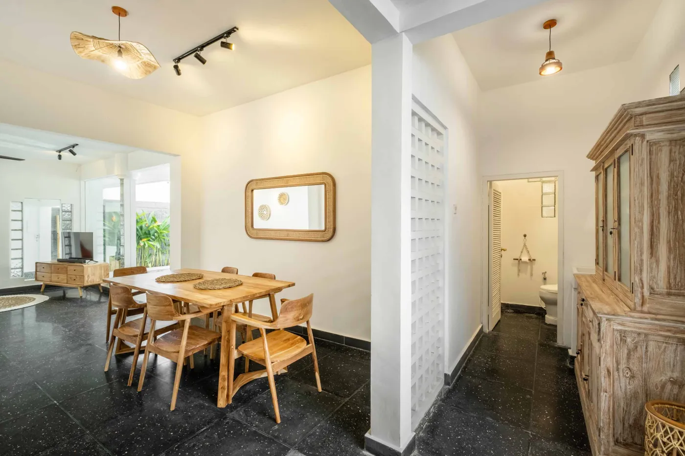 Modern tropical bathroom with dark tiled walls, rattan shelf and bamboo accents, oval rope mirror, white sink, bidet toilet, and window view of poolside greenery. - Bali Villas