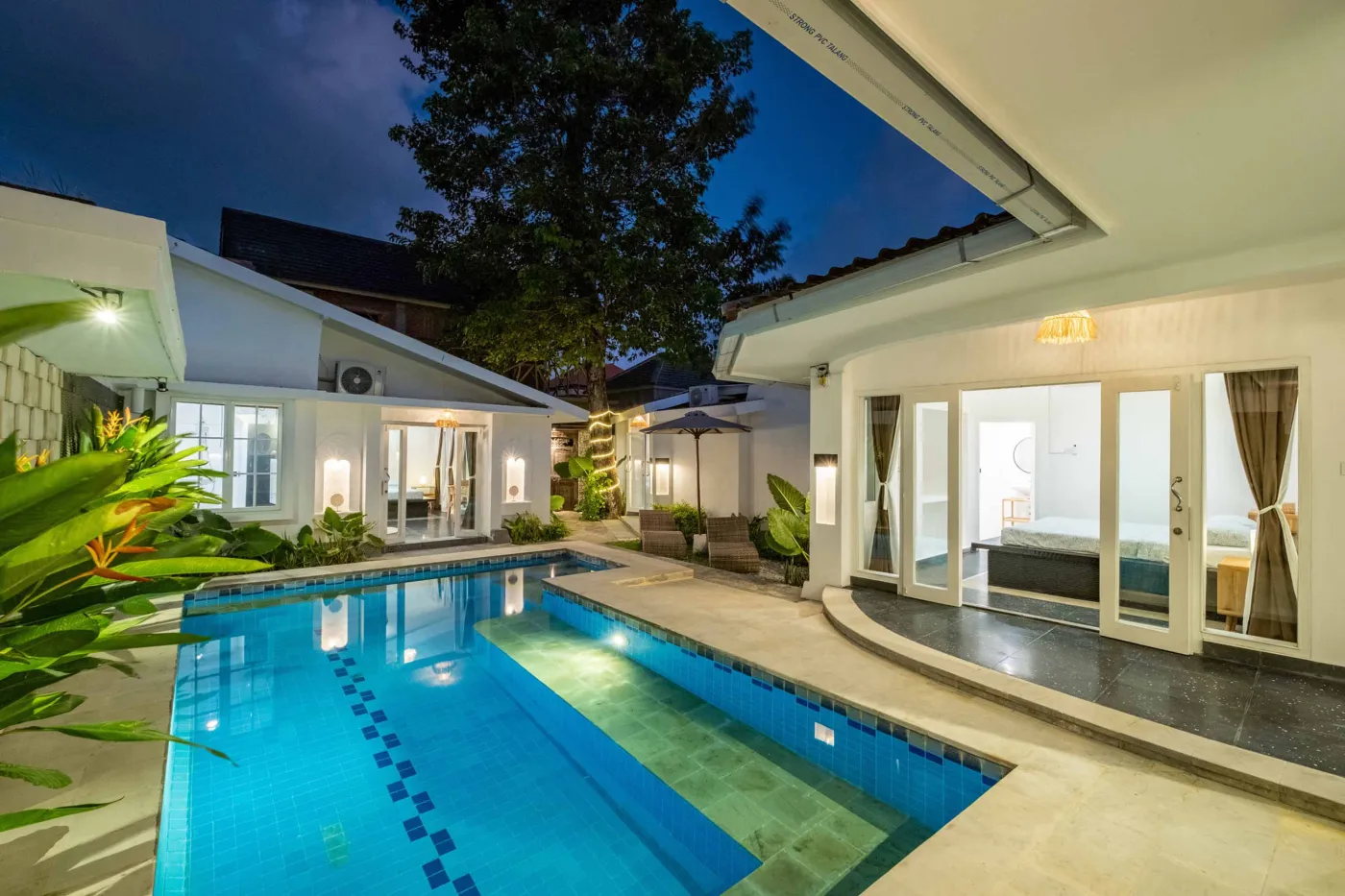 Modern tropical bathroom with dark tiled walls, rattan shelf and bamboo accents, oval rope mirror, white sink, bidet toilet, and window view of poolside greenery. - Bali Villas