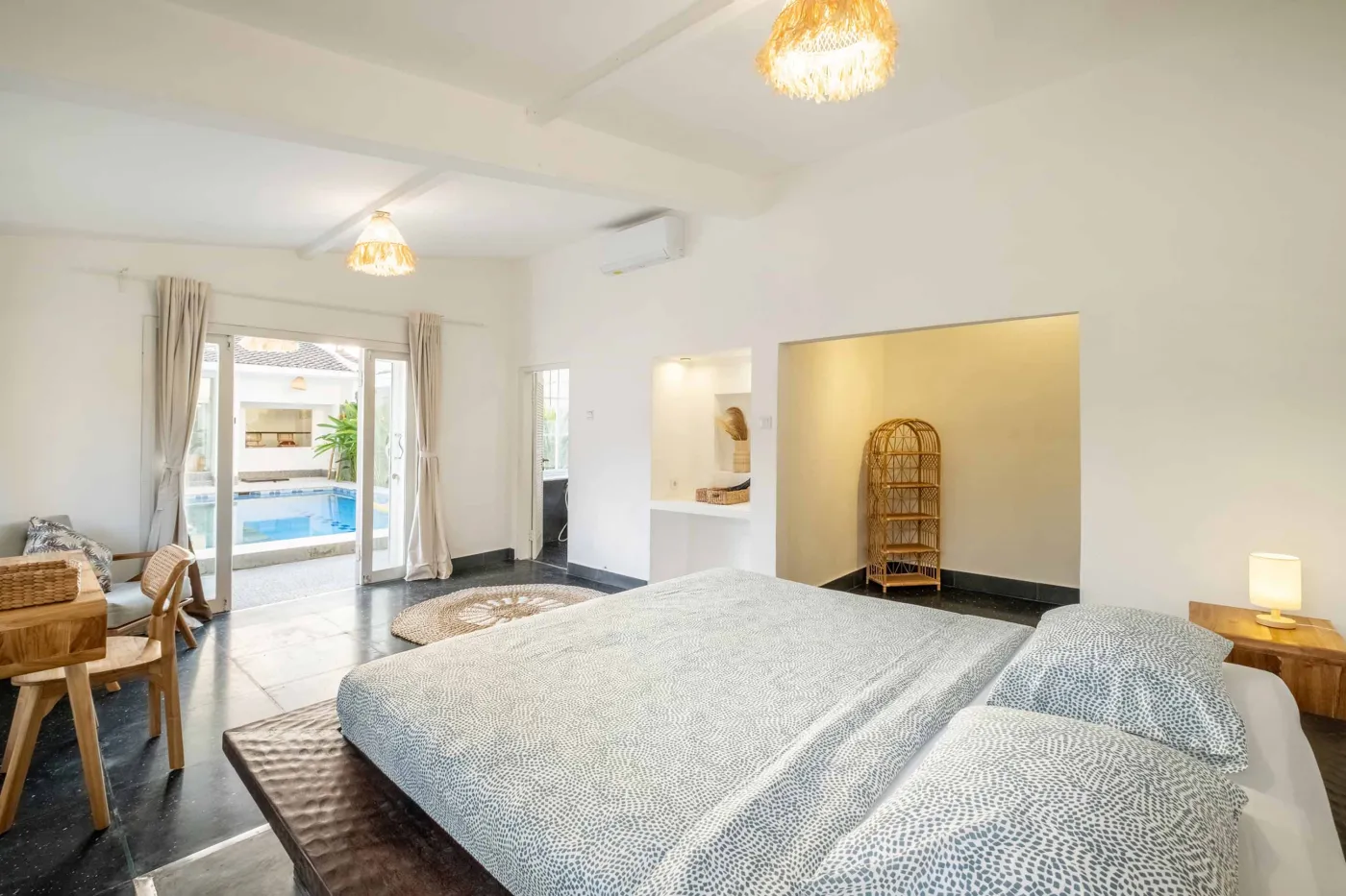 Modern tropical bathroom with dark tiled walls, rattan shelf and bamboo accents, oval rope mirror, white sink, bidet toilet, and window view of poolside greenery. - Bali Villas