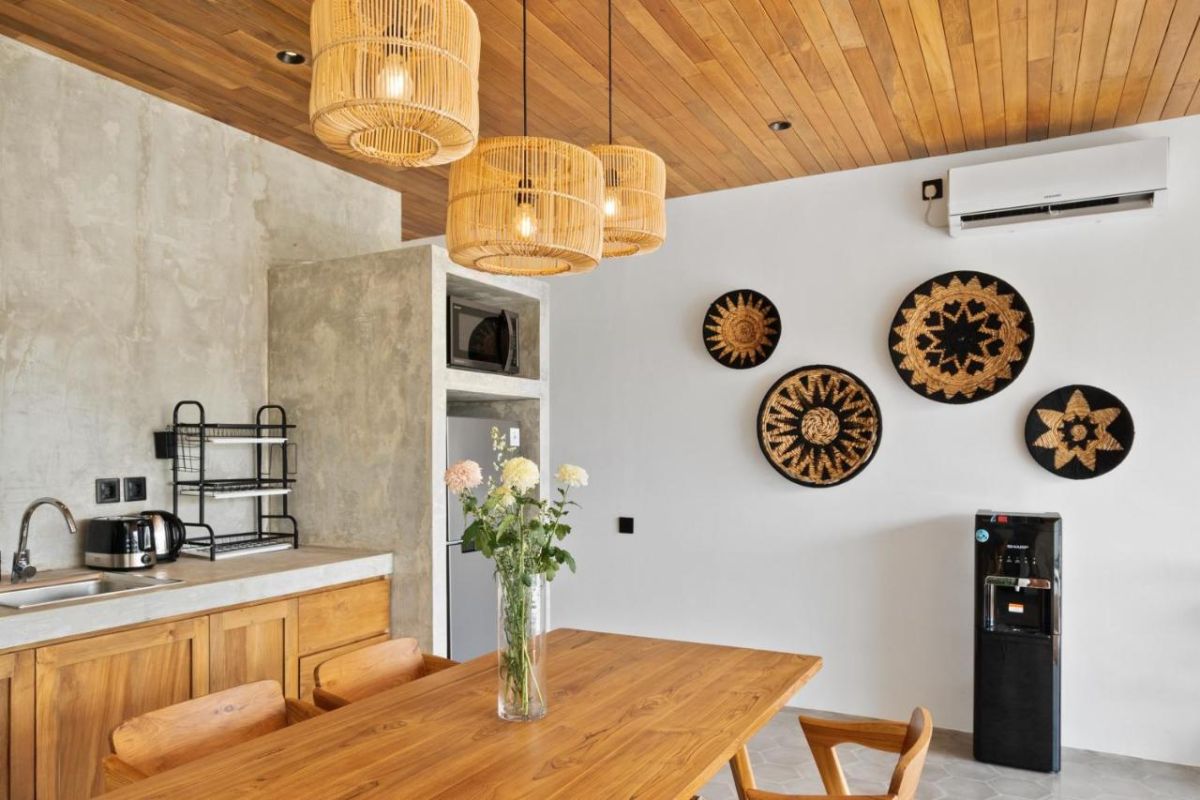 Modern tropical kitchen interior with wooden dining table, rattan pendant lights, woven wall art, and fresh flowers. - Bali Villas