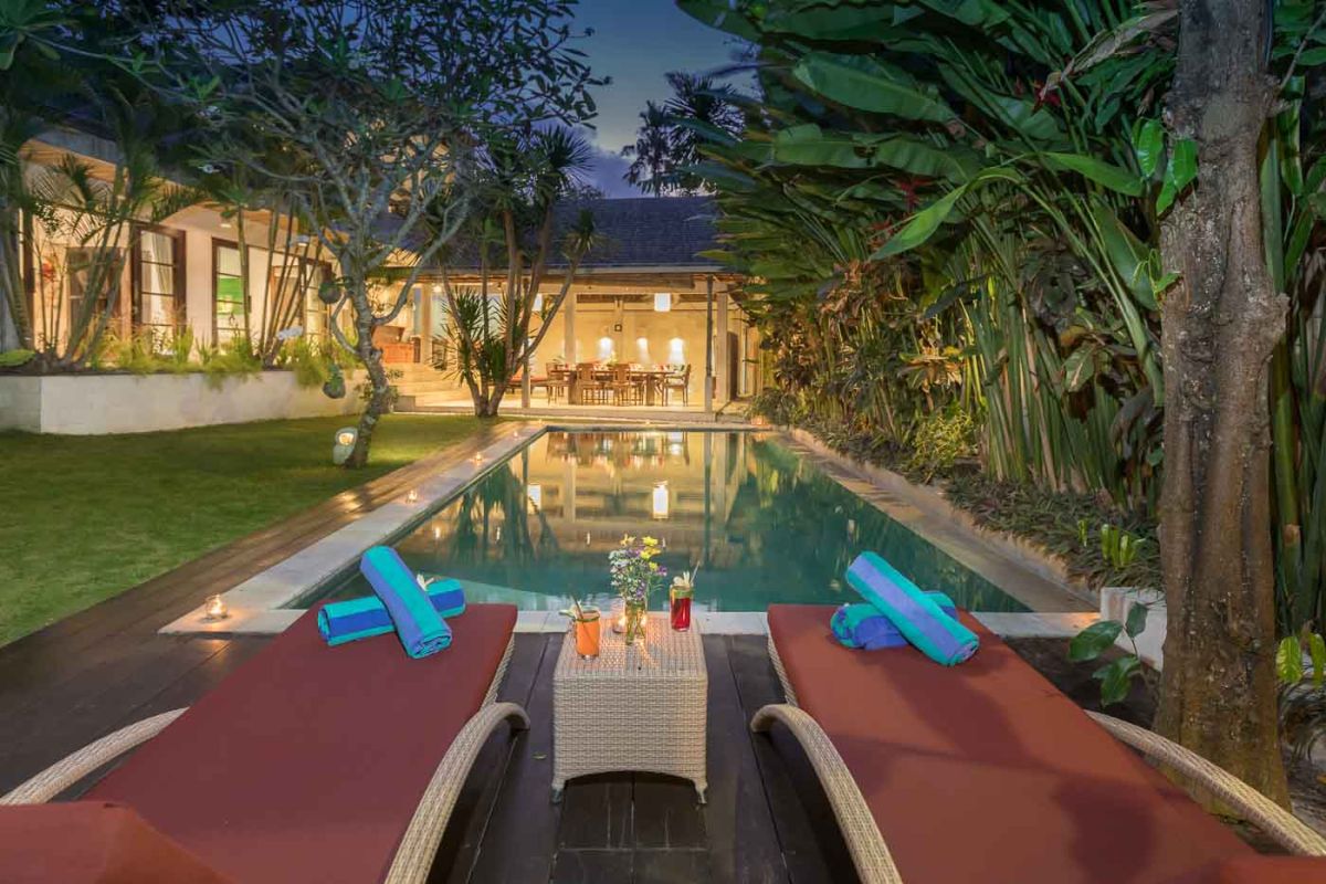 Spacious Balinese villa living room with teak wooden furniture, orange sofas, pendant lamps, and open views to tropical pool and garden. - Bali Villas