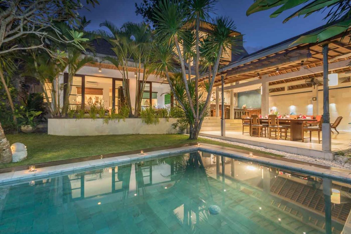 Spacious Balinese villa living room with teak wooden furniture, orange sofas, pendant lamps, and open views to tropical pool and garden. - Bali Villas