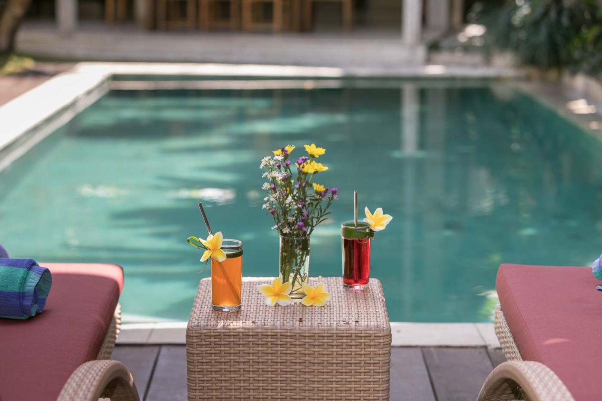 Spacious Balinese villa living room with teak wooden furniture, orange sofas, pendant lamps, and open views to tropical pool and garden. - Bali Villas