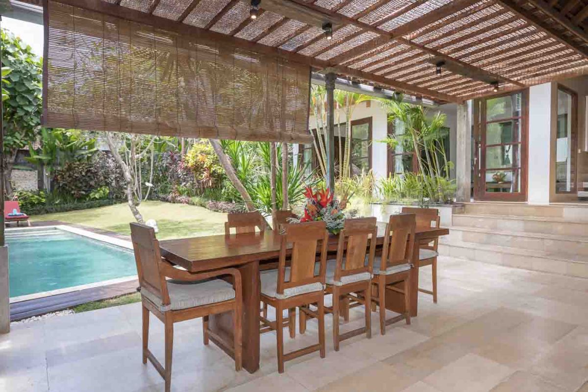 Spacious Balinese villa living room with teak wooden furniture, orange sofas, pendant lamps, and open views to tropical pool and garden. - Bali Villas