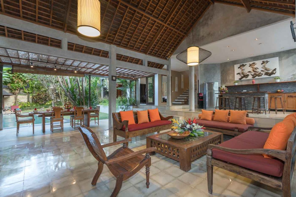 Spacious Balinese villa living room with teak wooden furniture, orange sofas, pendant lamps, and open views to tropical pool and garden. - Bali Villas