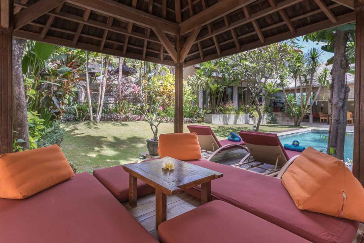 Spacious Balinese villa living room with teak wooden furniture, orange sofas, pendant lamps, and open views to tropical pool and garden. - Bali Villas