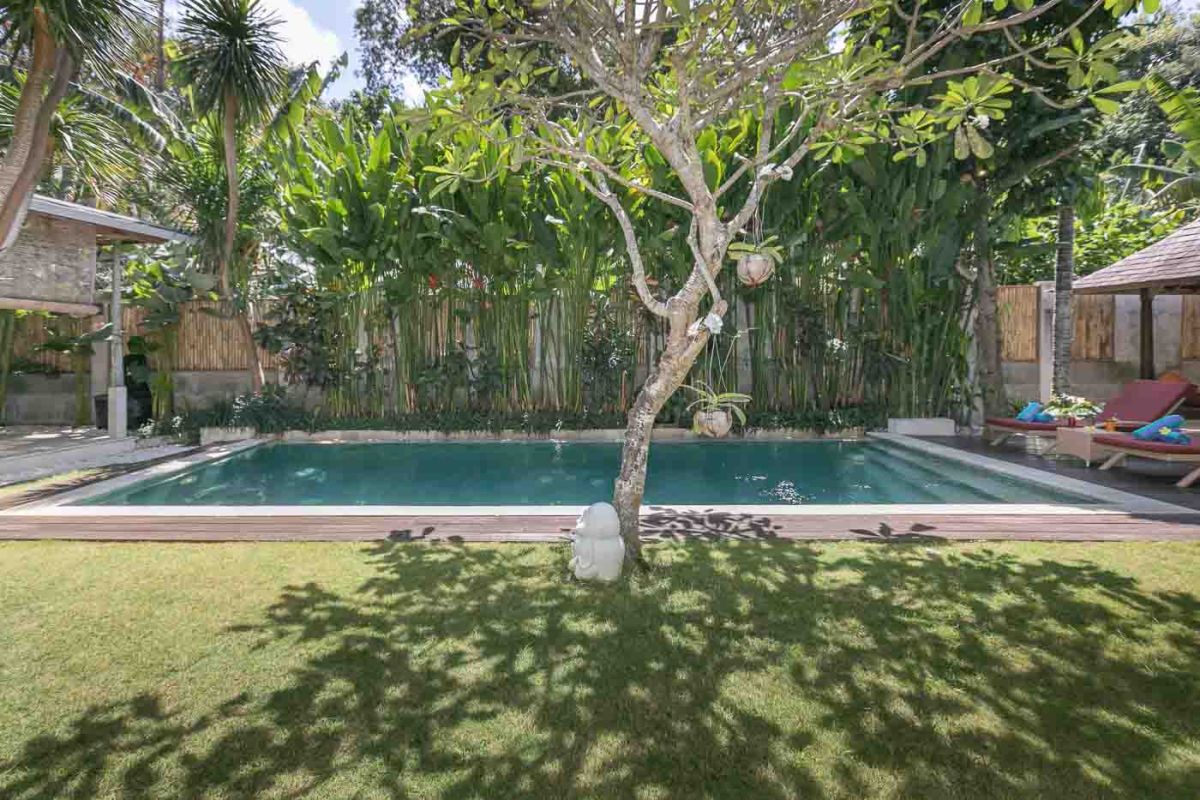 Spacious Balinese villa living room with teak wooden furniture, orange sofas, pendant lamps, and open views to tropical pool and garden. - Bali Villas