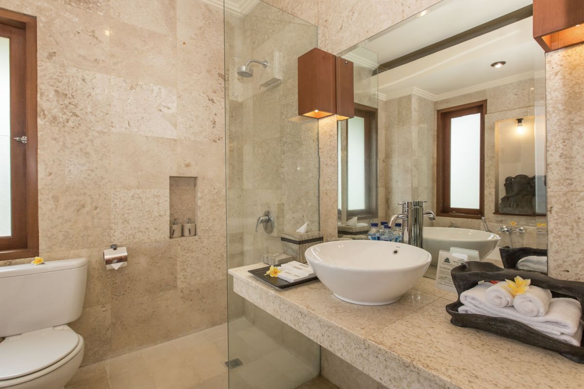 Modern luxury bathroom with beige stone tiles, freestanding white bathtub, glass shower enclosure, and wooden accents. - Bali Villas