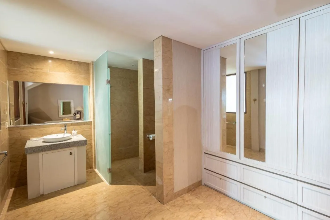 Modern en-suite bathroom featuring white vanity sink, glass shower enclosure, and adjacent mirrored wardrobe on beige tiled floor. - Bali Villas