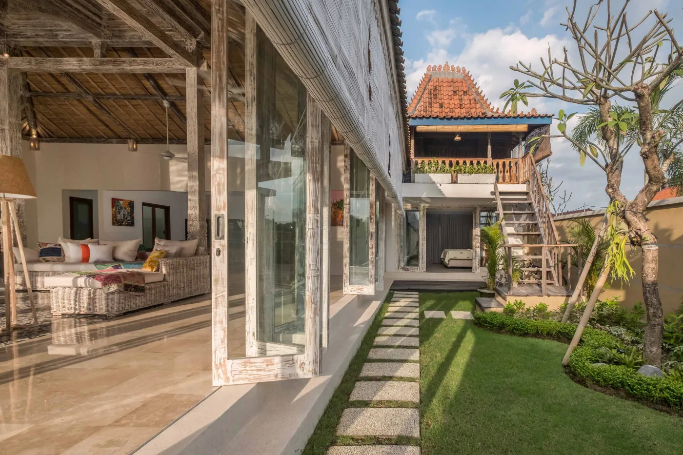 Modern Balinese villa with open-plan living room featuring white sofa, large glass doors to lush garden path, wooden accents, and red-tiled upper level. - Bali Villas