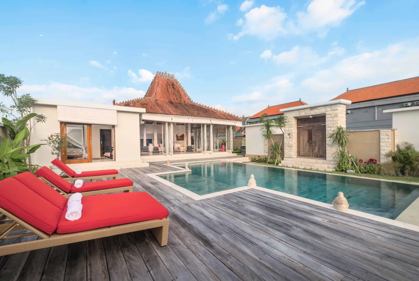 Modern Balinese villa with open-plan living room featuring white sofa, large glass doors to lush garden path, wooden accents, and red-tiled upper level. - Bali Villas