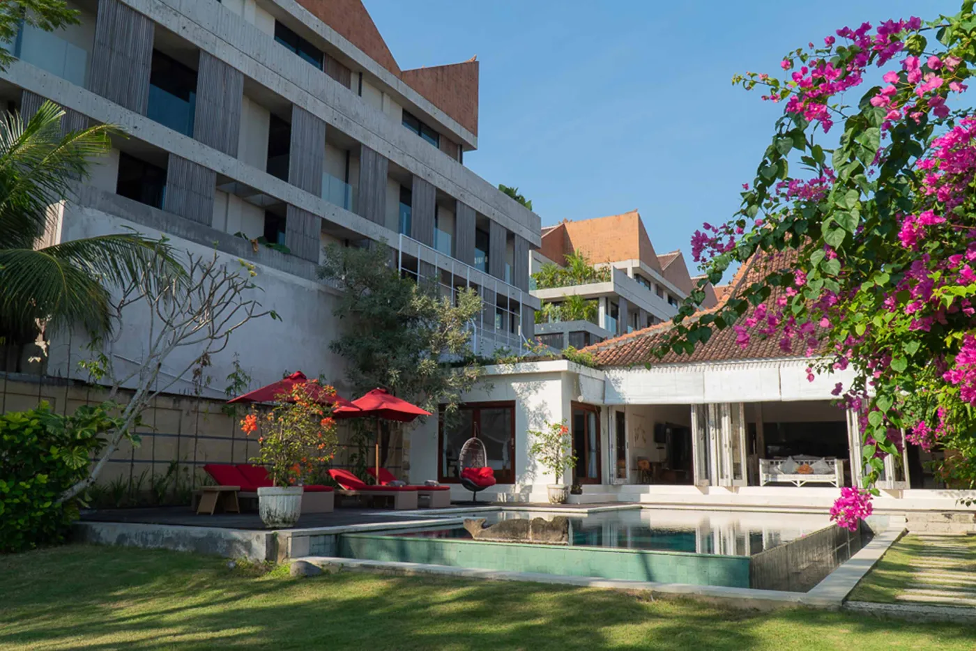 Modern Balinese villa with open-plan living room featuring white sofa, large glass doors to lush garden path, wooden accents, and red-tiled upper level. - Bali Villas