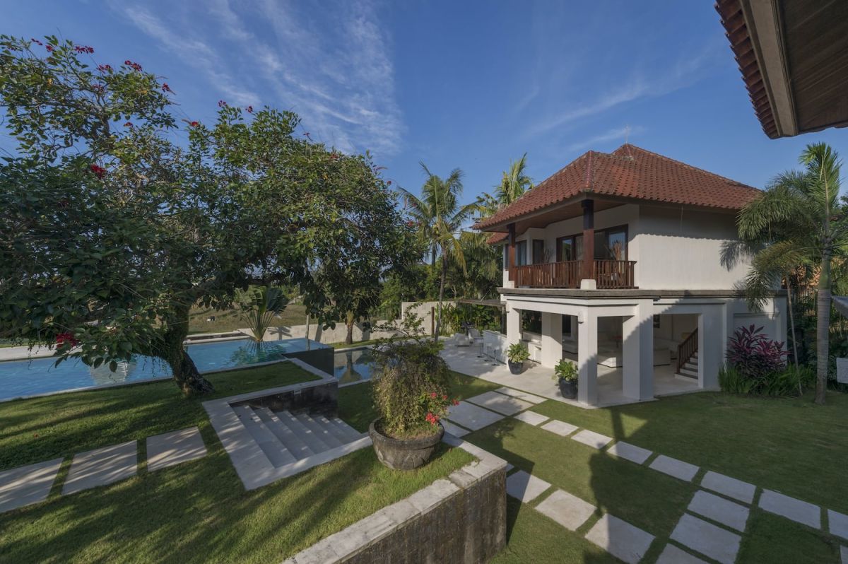 Modern white Bali villa with red-tiled roof, wooden balcony, infinity pool, and lush tropical garden under blue sky. - Bali Villas