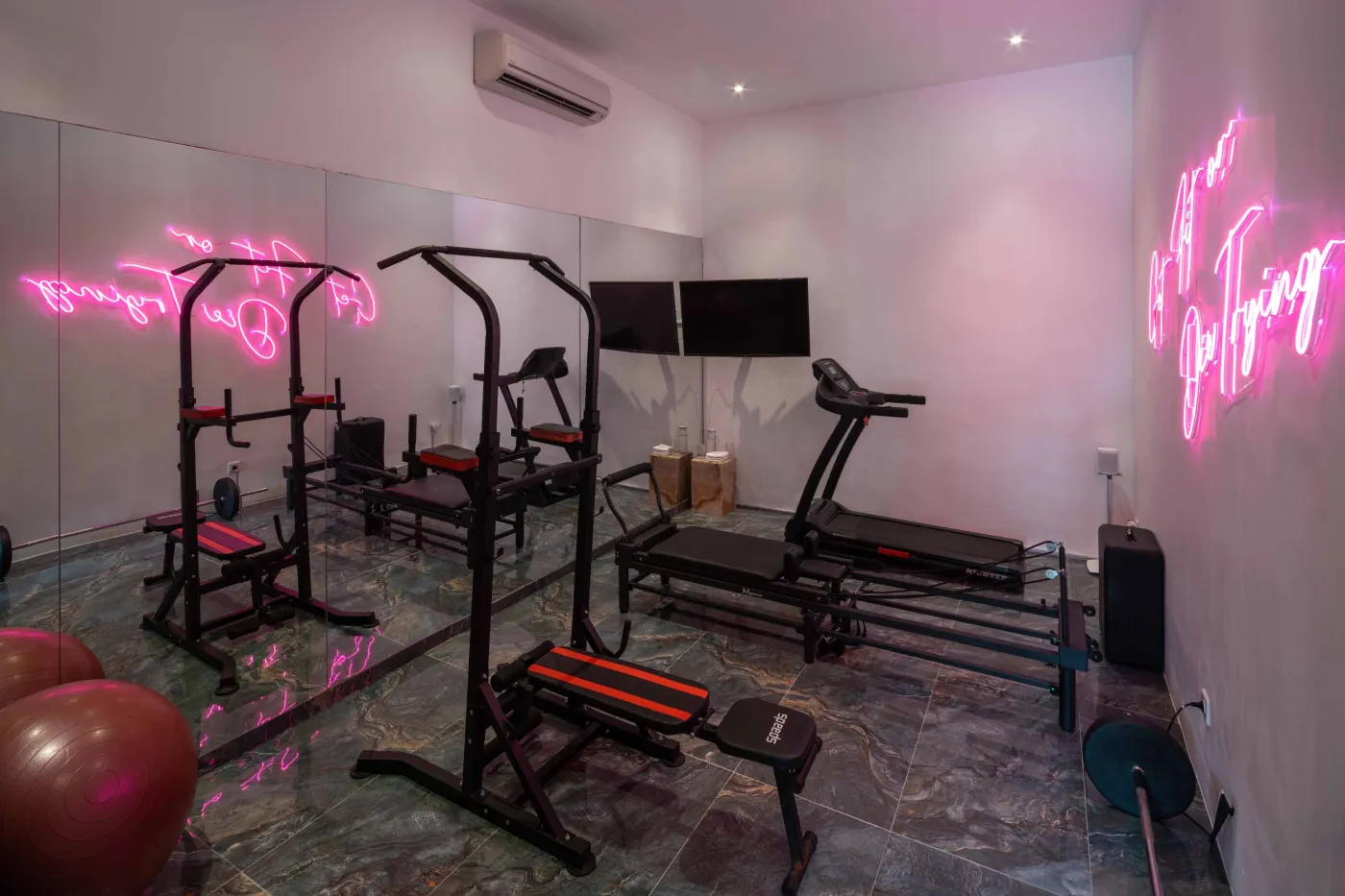 Modern home gym featuring a treadmill, weight bench, pull-up bars, exercise balls, and pink "Lifting" neon sign in a mirrored room. - Bali Villas