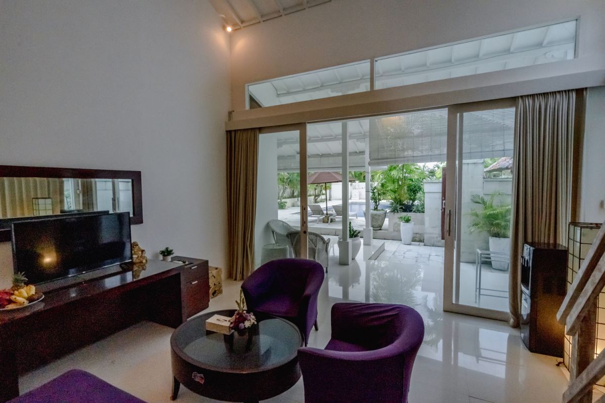 Spacious Balinese loft living room with wooden mezzanine stairs, purple sofa, TV console, white coffee table, and tropical wall art. - Bali Villas