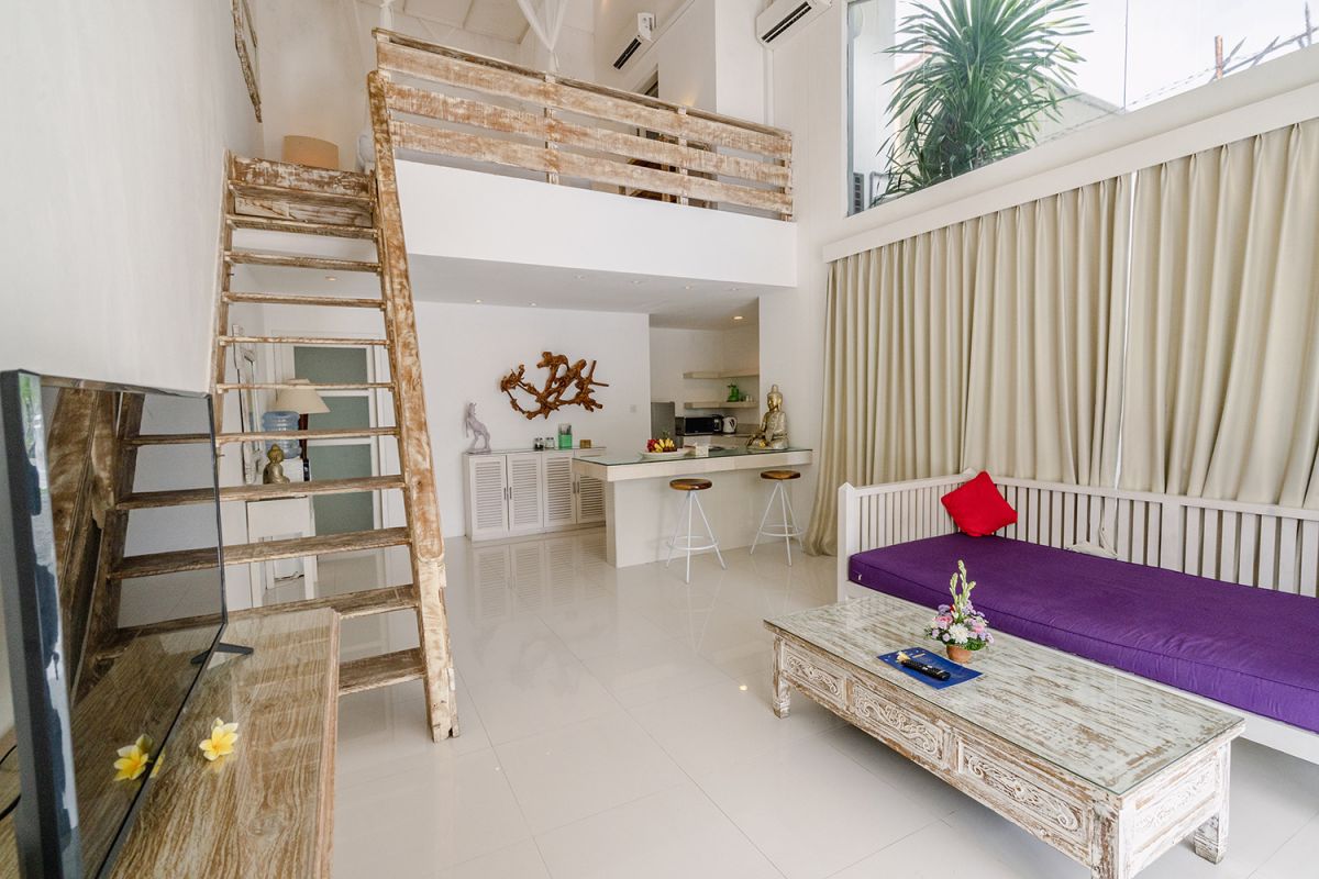 Spacious Balinese loft living room with wooden mezzanine stairs, purple sofa, TV console, white coffee table, and tropical wall art. - Bali Villas