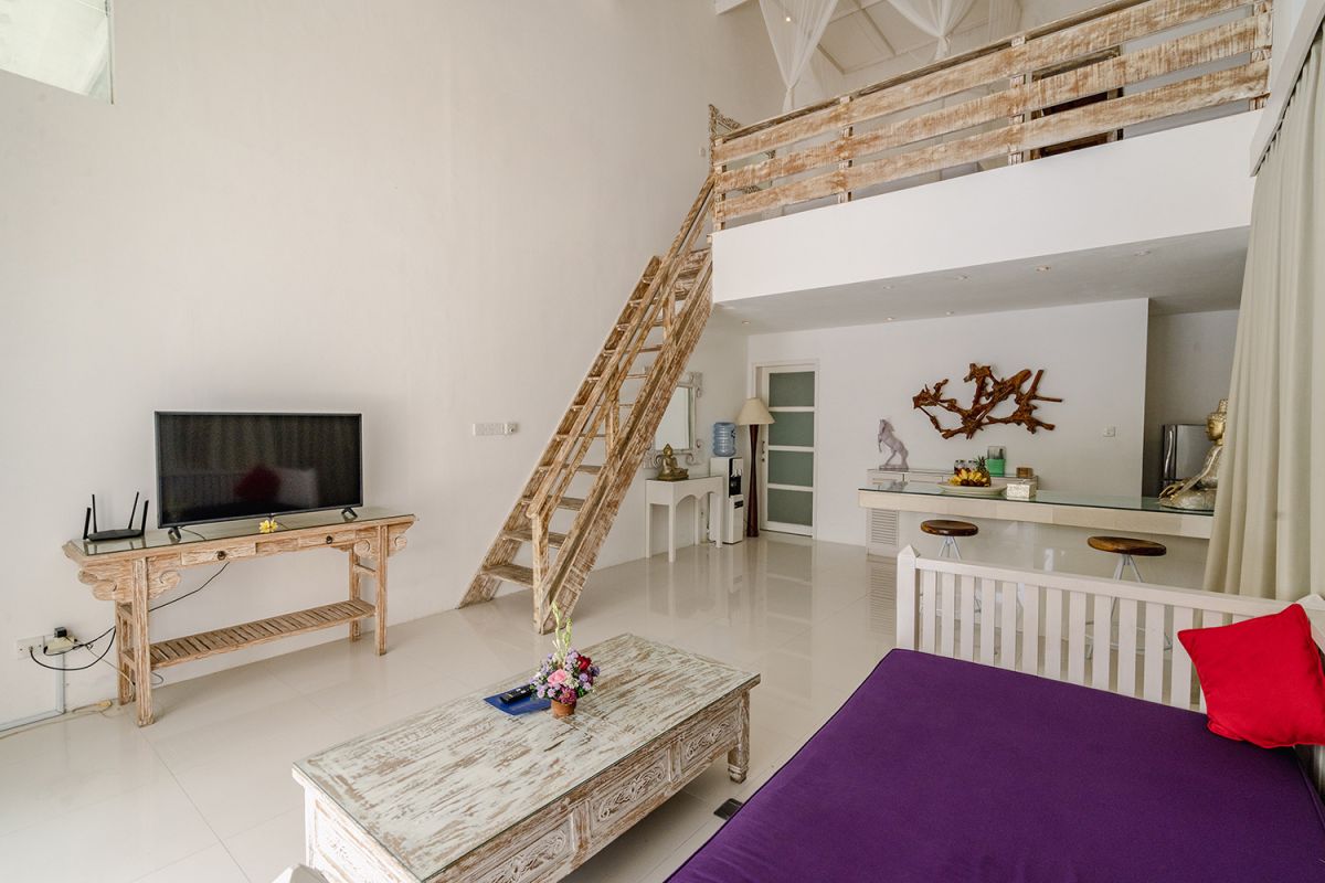 Spacious Balinese loft living room with wooden mezzanine stairs, purple sofa, TV console, white coffee table, and tropical wall art. - Bali Villas