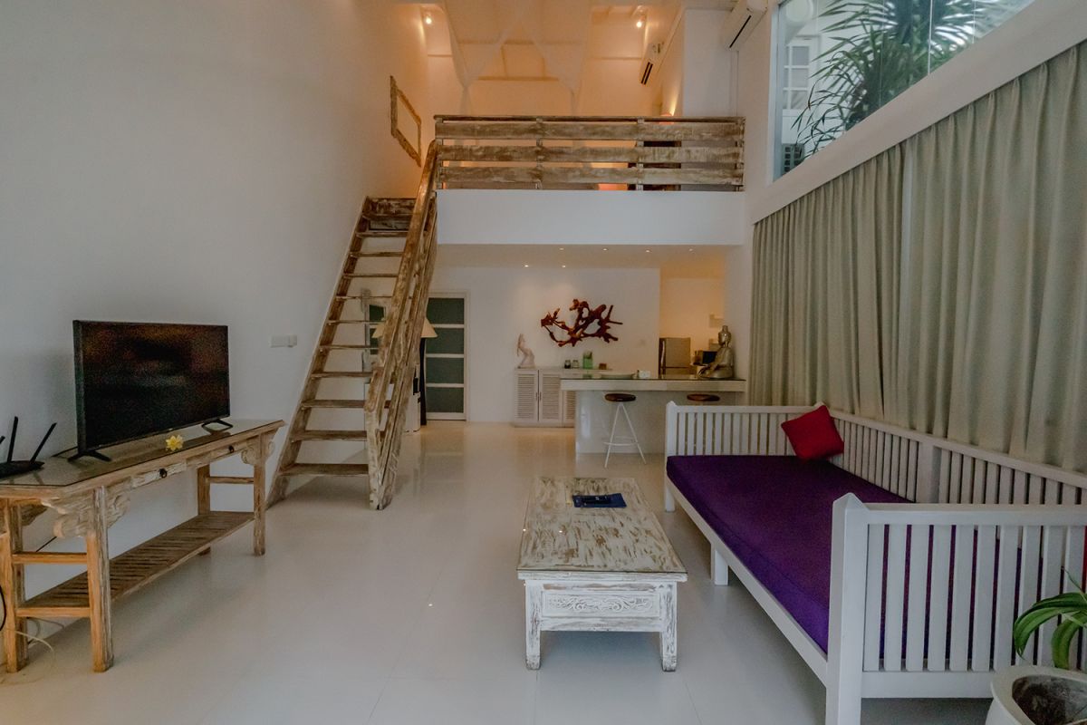 Spacious Balinese loft living room with wooden mezzanine stairs, purple sofa, TV console, white coffee table, and tropical wall art. - Bali Villas