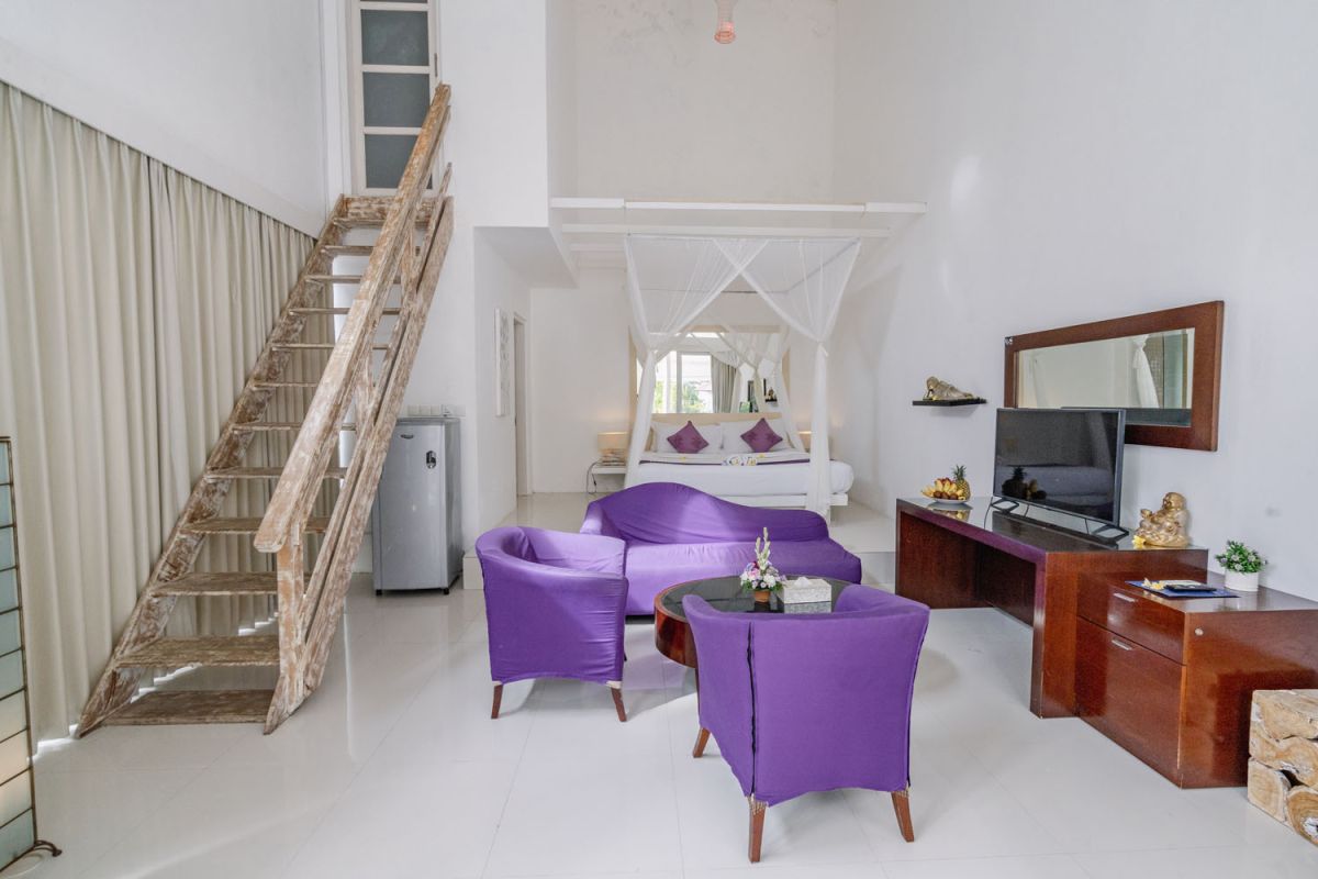 Spacious Balinese loft living room with wooden mezzanine stairs, purple sofa, TV console, white coffee table, and tropical wall art. - Bali Villas
