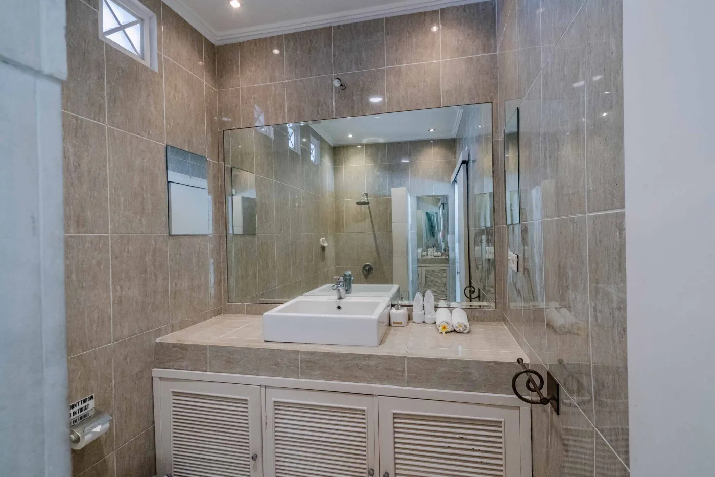 Modern bathroom interior with beige tiled walls, large frameless mirror, white vanity sink, toiletries, and walk-in shower. - Bali Villas