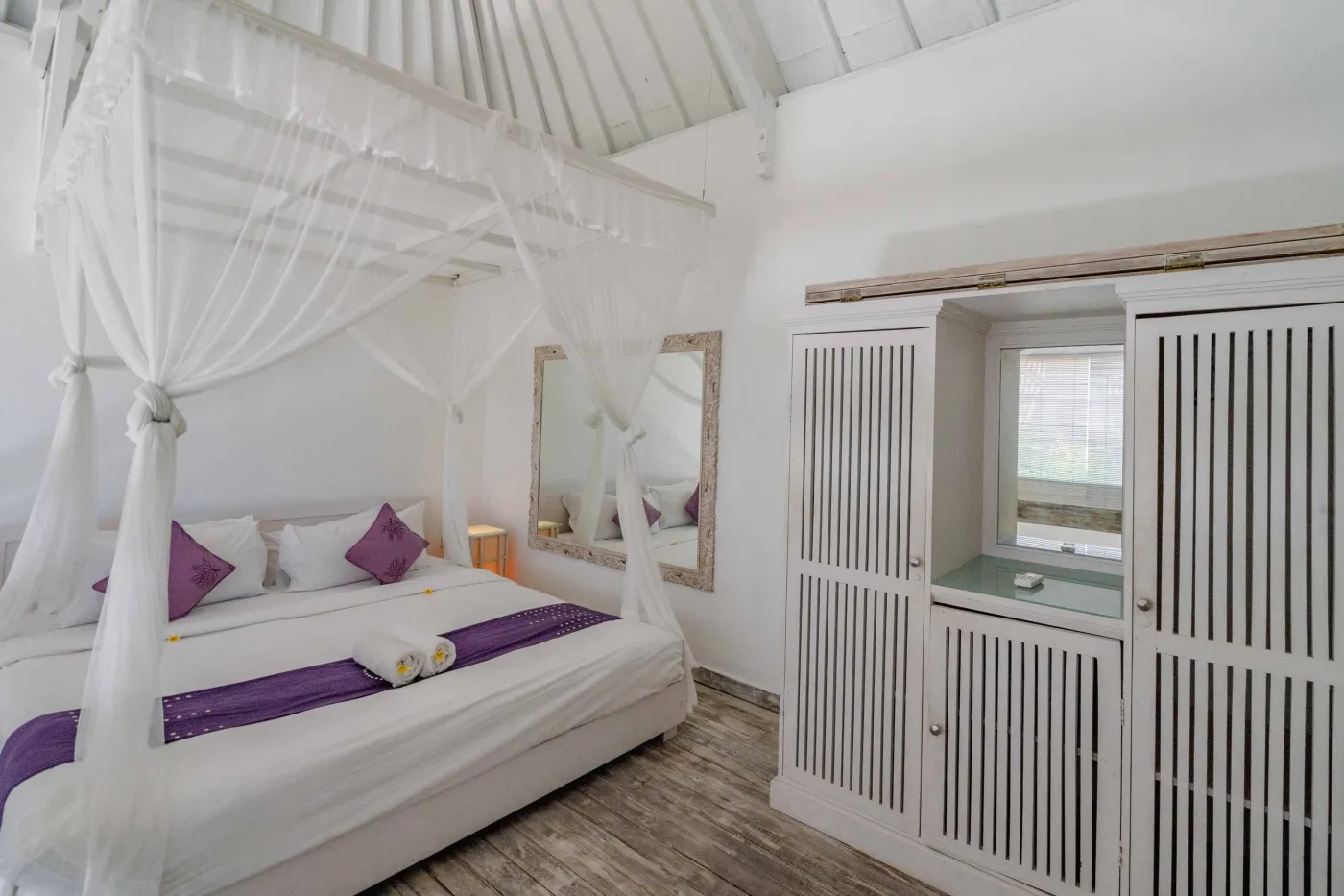 Serene Balinese bedroom with white canopy four-poster bed, purple pillows, and louvered wooden wardrobe in a thatched-roof villa. - Bali Villas