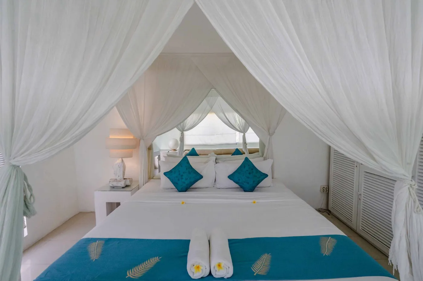 Modern open-plan tropical interior with sliding glass doors connecting living room to outdoor seating area, white tiled floors, plush sofas, and potted plants. - Bali Villas