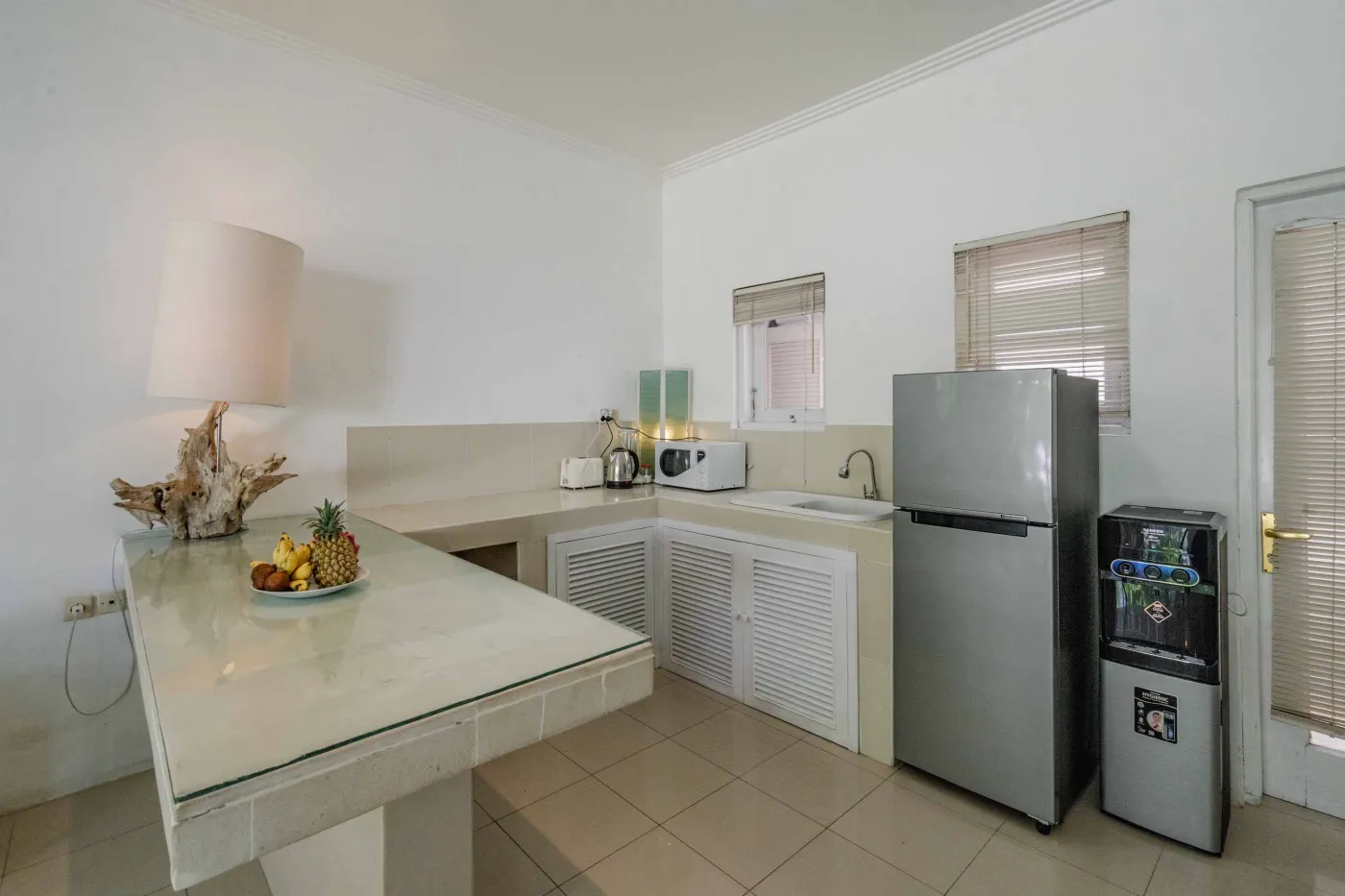 Modern open-plan tropical interior with sliding glass doors connecting living room to outdoor seating area, white tiled floors, plush sofas, and potted plants. - Bali Villas
