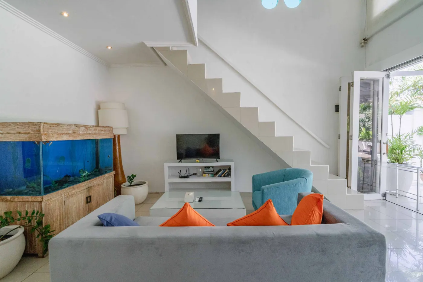 Modern open-plan tropical interior with sliding glass doors connecting living room to outdoor seating area, white tiled floors, plush sofas, and potted plants. - Bali Villas