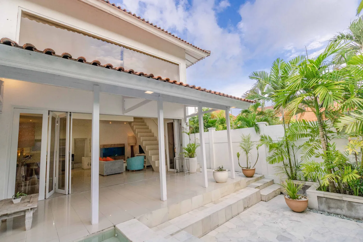 Modern open-plan tropical interior with sliding glass doors connecting living room to outdoor seating area, white tiled floors, plush sofas, and potted plants. - Bali Villas