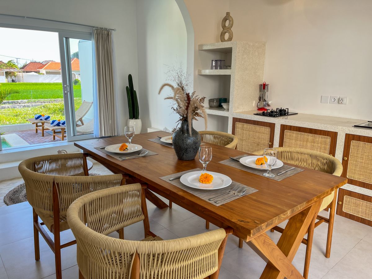 Elegant Bali villa dining room with wooden table set for meal, rattan chairs, and tropical pool view through glass doors. - Bali Villas
