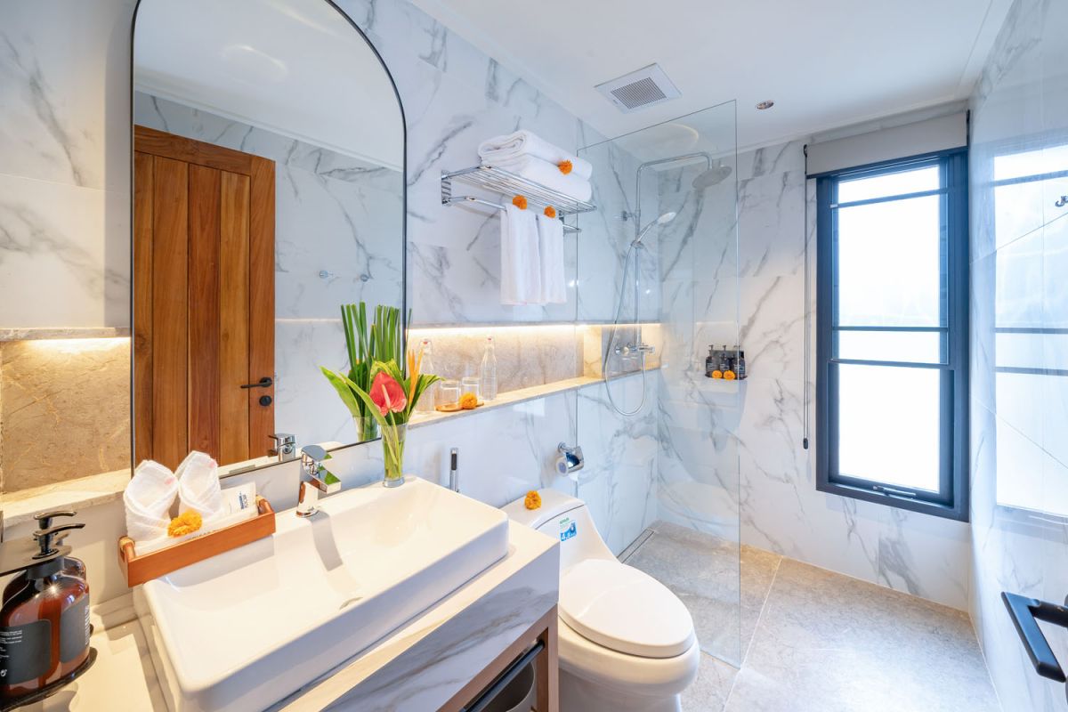 Modern luxury bathroom featuring white marble walls, wooden vanity sink, arched mirror, tropical red flower arrangement, and glass-enclosed shower. - Bali Villas