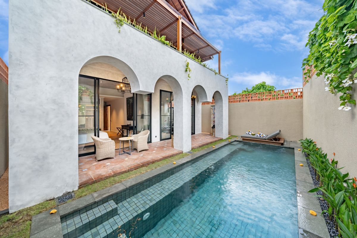 Modern luxury bathroom featuring white marble walls, wooden vanity sink, arched mirror, tropical red flower arrangement, and glass-enclosed shower. - Bali Villas