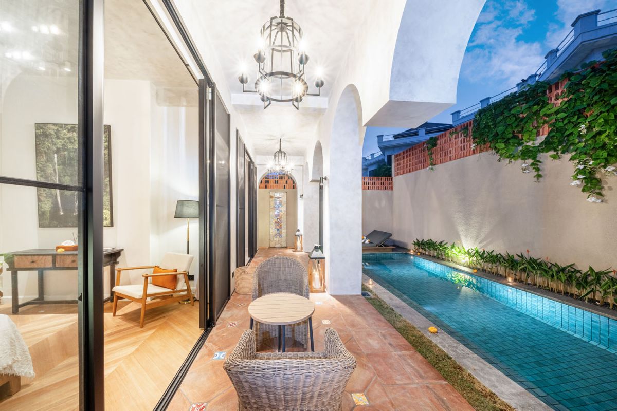 Elegant arched hallway with wooden floors, modern furniture, and glass doors opening to a narrow lit poolside garden at dusk. - Bali Villas