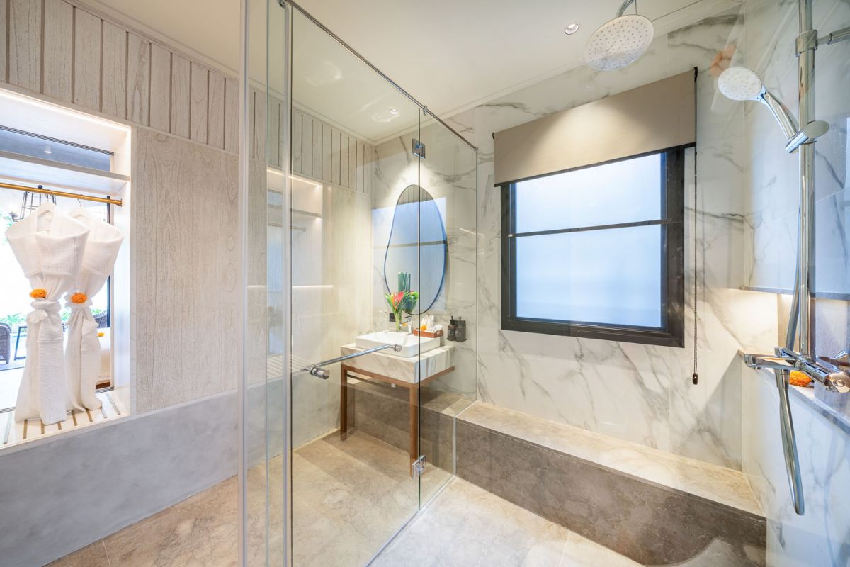 Modern luxury bathroom featuring white marble walls, wooden vanity sink, arched mirror, tropical red flower arrangement, and glass-enclosed shower. - Bali Villas