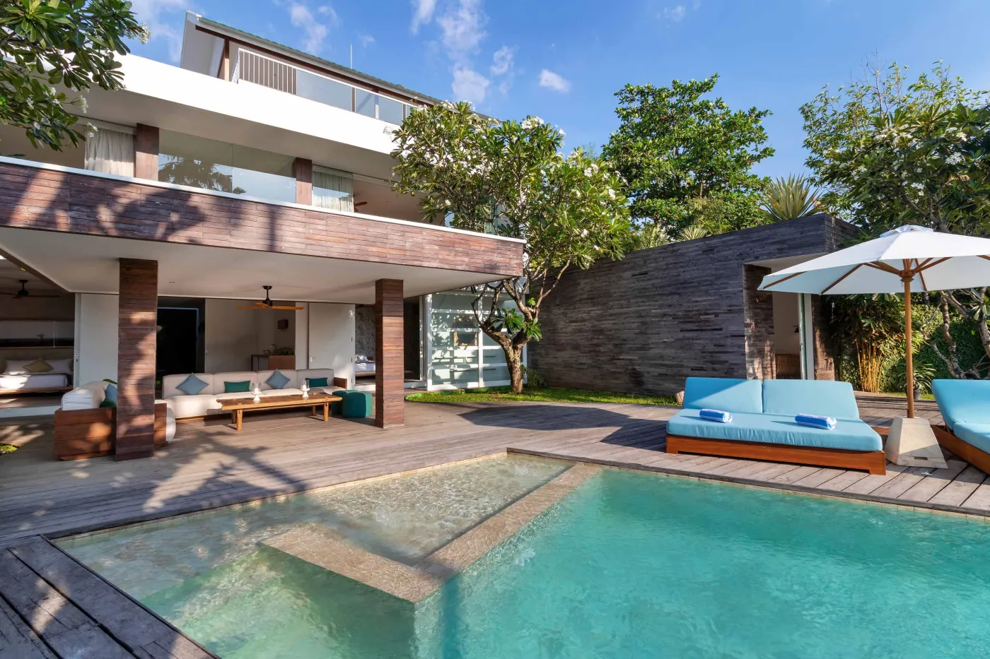 Modern tropical villa with infinity-edge pool, wooden deck lounge area, and lush greenery under clear skies. - Bali Villas