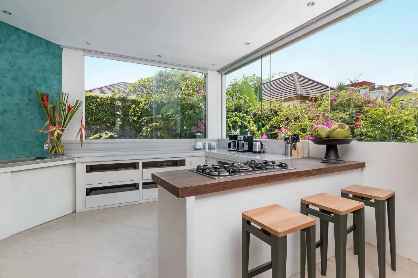 Cozy outdoor balcony lounge with white armchairs, blue tiled table, and potted plants, overlooking tropical palm trees at dusk sunset. - Bali Villas
