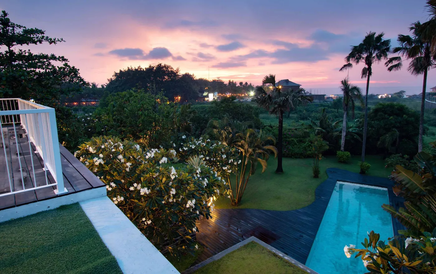 Cozy outdoor balcony lounge with white armchairs, blue tiled table, and potted plants, overlooking tropical palm trees at dusk sunset. - Bali Villas