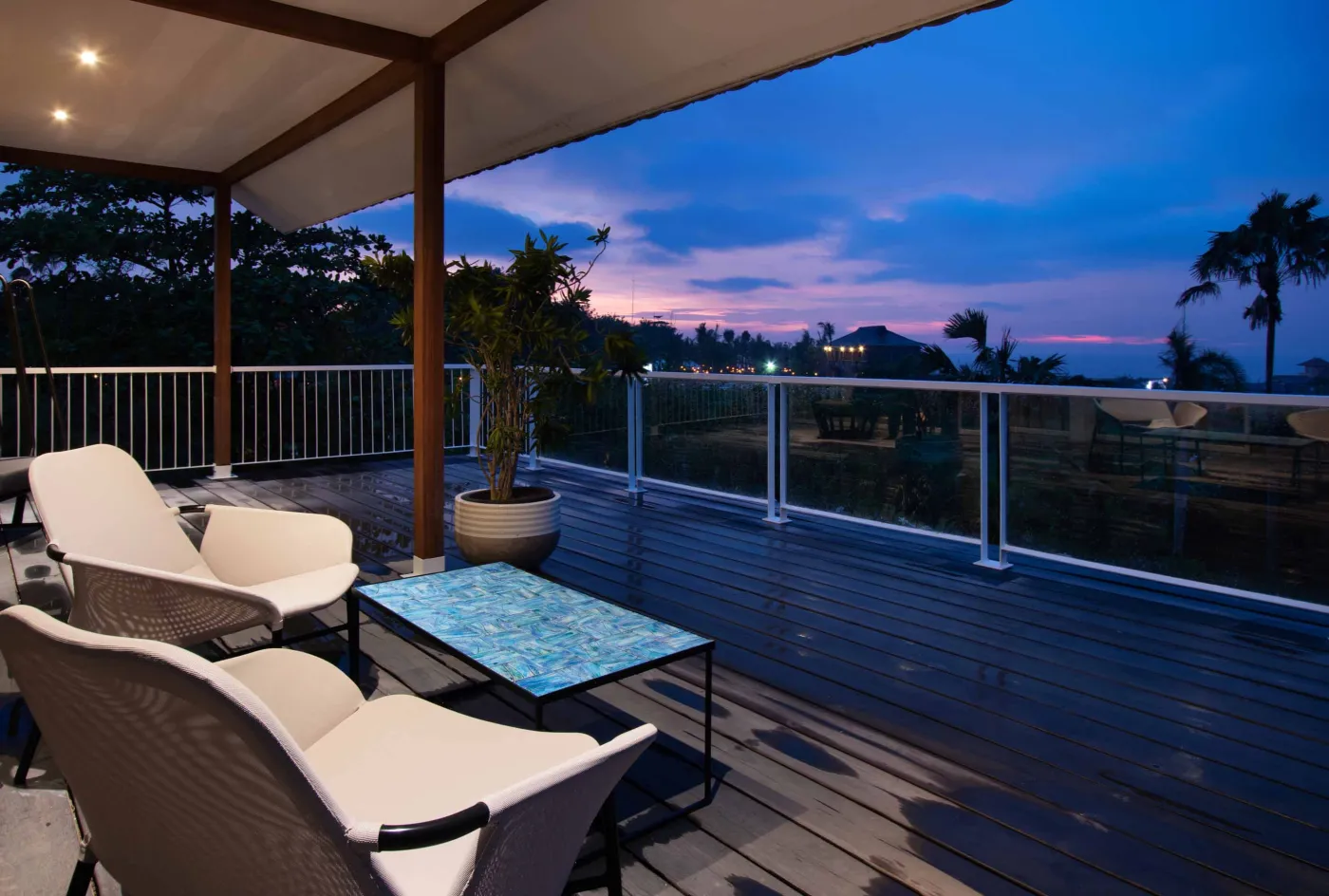 Cozy outdoor balcony lounge with white armchairs, blue tiled table, and potted plants, overlooking tropical palm trees at dusk sunset. - Bali Villas