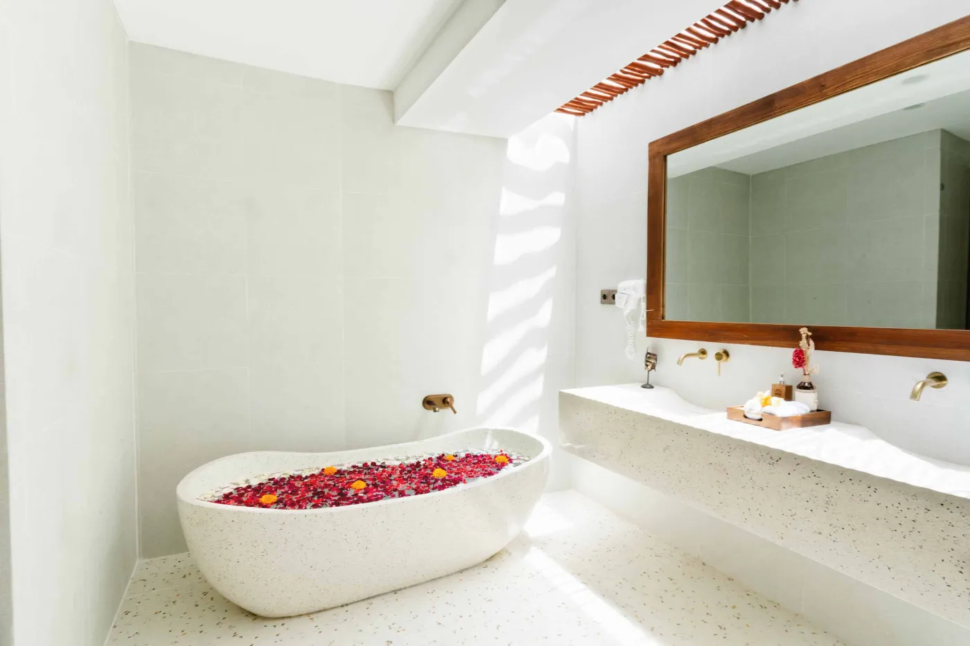Luxurious spa bathroom with white freestanding bathtub filled with floating red hibiscus petals, wooden mirror, and brass fixtures in soft natural light. - Bali Villas