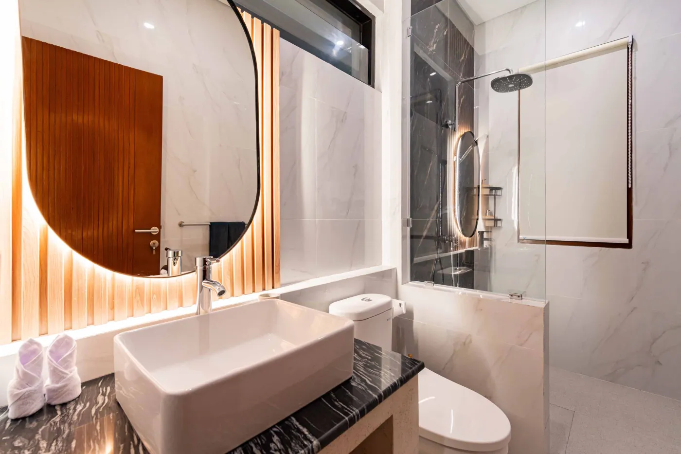 Modern bathroom with round wooden-framed mirror, white vessel sink on black countertop, toilet, and glass shower enclosure. - Bali Villas