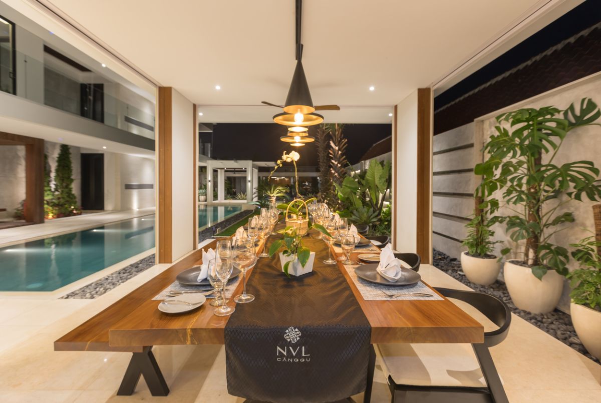 Elegant outdoor dining table set for dinner in a luxurious Bali villa, with tropical plants, pool view, and NVYL branding at night. - Bali Villas