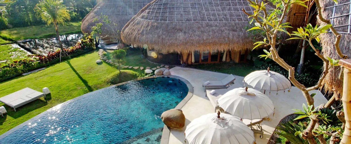 Spacious open-plan Bali villa living area with white sofas, rustic wooden coffee table, bamboo ceiling, tropical plants, and Buddha statue. - Bali Villas