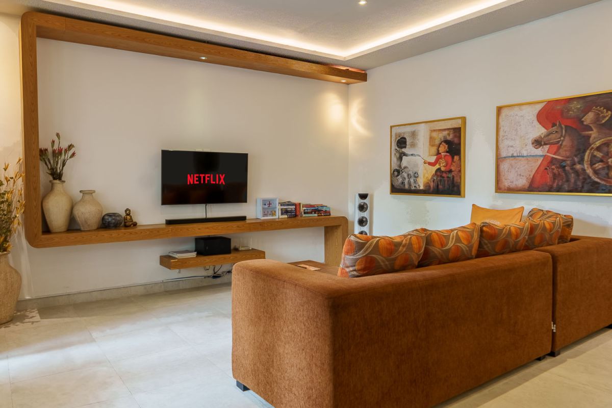 Modern living room with wooden wall panels, orange patterned sofa, Netflix on wall-mounted TV, abstract wall art, and potted plants. - Bali Villas