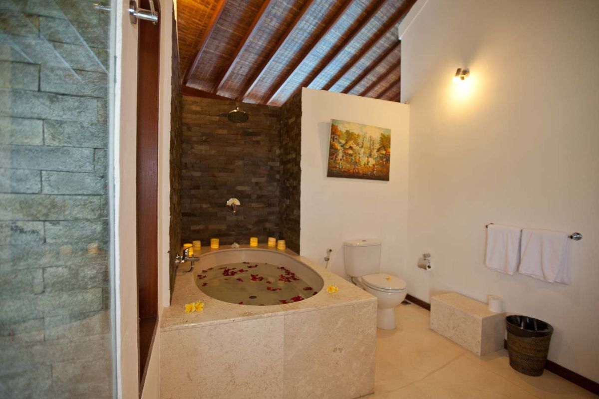 Luxurious Balinese bathroom with petal-filled stone bathtub, rain shower, wooden beamed ceiling, and tropical accents. - Bali Villas