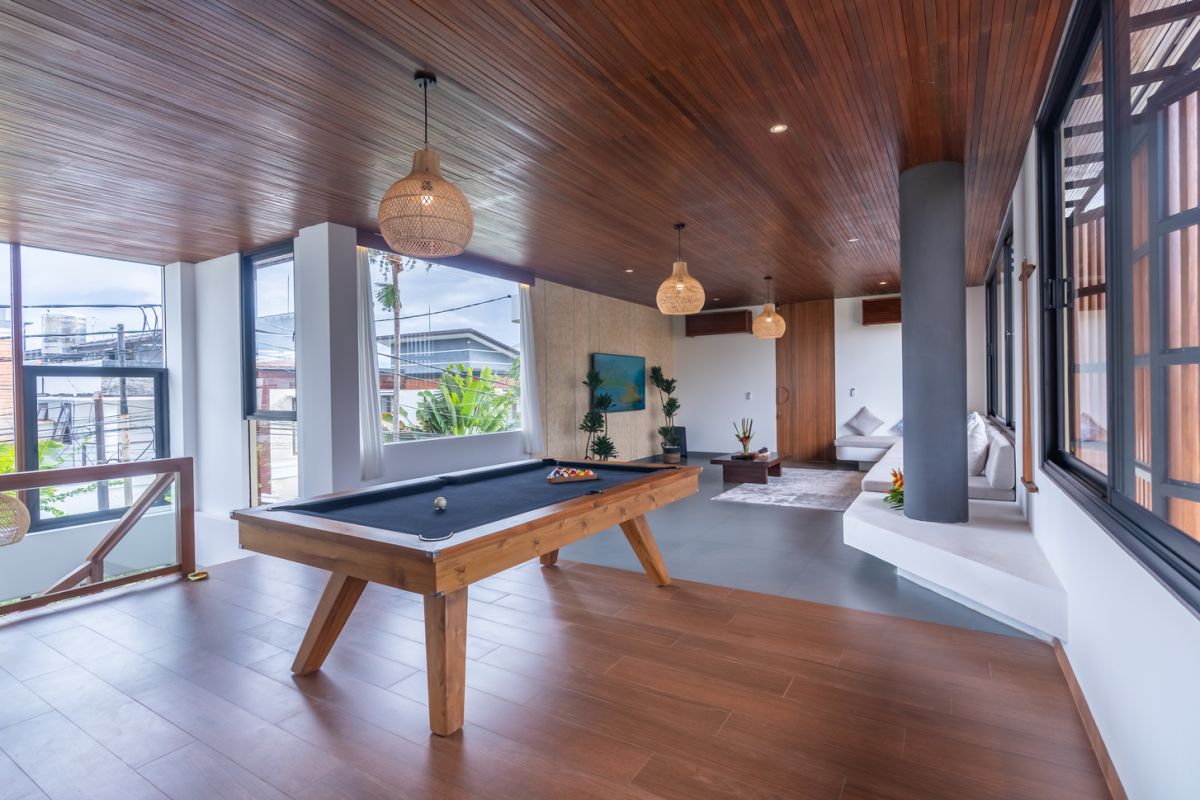 Modern wooden pool table in a spacious tropical living room with large windows, plants, and cozy seating. - Bali Villas