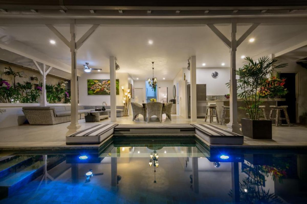 Luxurious open-air Balinese villa featuring an infinity pool, gray lounge seating, wooden dining table, and tropical plants under a blue sky. - Bali Villas